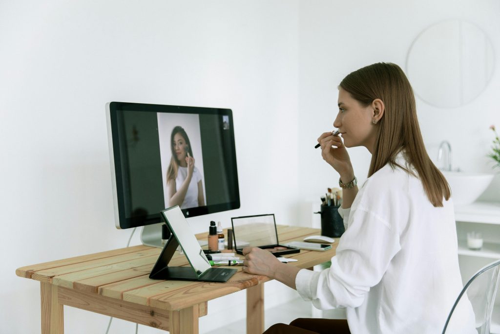 Woman following an online makeup tutorial, applying cosmetics, sitting at a wooden desk.